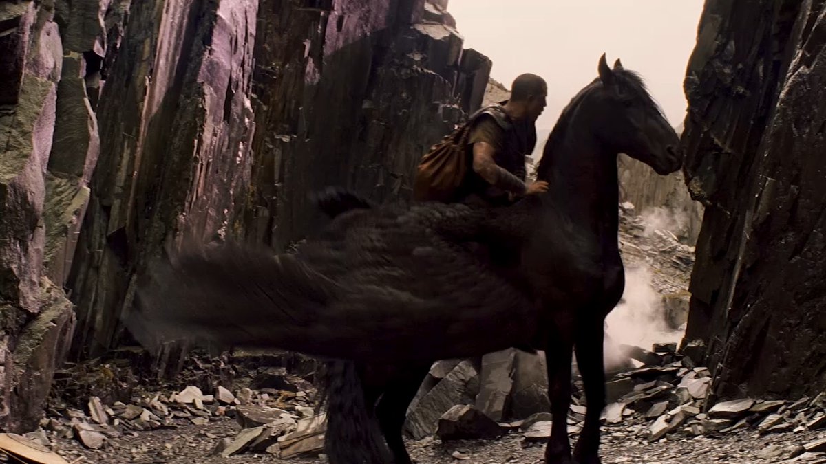 A warrior rides a black horse through a rocky, smoke-filled canyon, evoking the epic journey described in the promotional post about facing terrifying creatures in a war between the gods. The scene captures the adventurous and mythological tone of "Clash of the Titans" and its sequel, airing Saturday at 2:00 PM MEX on #El5.
