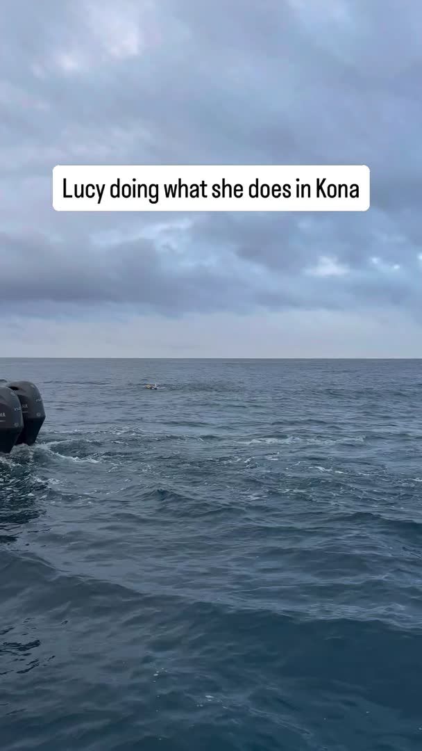 A view from a boat on a cloudy day with the caption "Lucy doing what she does in Kona", showing a small figure swimming in the ocean with a Yamaha motor visible on the left.
