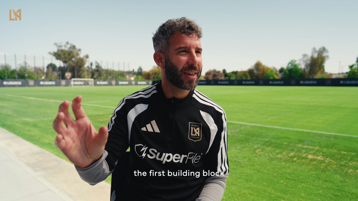 A bearded man in a black LAFC training jacket with "SuperFile" sponsorship speaks animatedly on a sunlit soccer training pitch, gesturing with his hand as the caption "the first building block" appears on screen—visually reinforcing the post's message of laying the groundwork for the mission ahead.