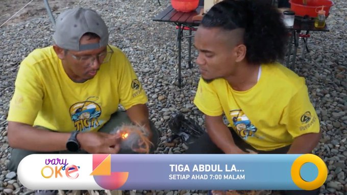 Two men in yellow "TIGA" shirts sit outdoors on gravel, with a TV overlay reading "vary OK" and "TIGA ABDUL LA... SETIAP AHAD 7:00 MALAM".