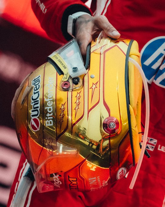 A close-up of a Ferrari racing helmet featuring reflective gold coloring, sponsor logos including "UniCredit" and "Bitdefender," and a clear drinking tube, held by a hand emerging from a red racing suit. The image captures the nostalgic, polished aesthetic of a Ferrari driver's gear, fitting the "Reminiscence of a Saturday Sprint" caption by evoking the glamour and tradition of Formula 1 race weekends.