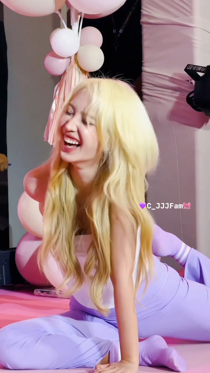 A woman with long blonde hair laughs joyfully while sitting on the floor in a pink and purple outfit, with pink balloons behind her and a 'C_JJJFam' watermark.