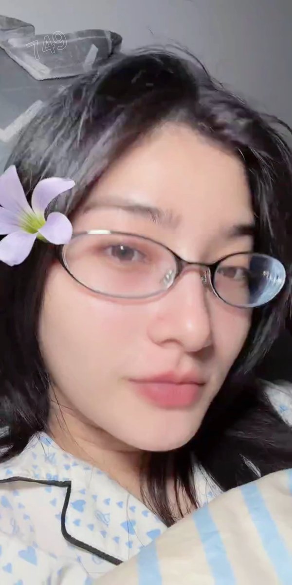 A close-up selfie of a woman with dark hair, round glasses, and a digital purple flower filter on her cheek; the number '749' appears in the upper left corner.