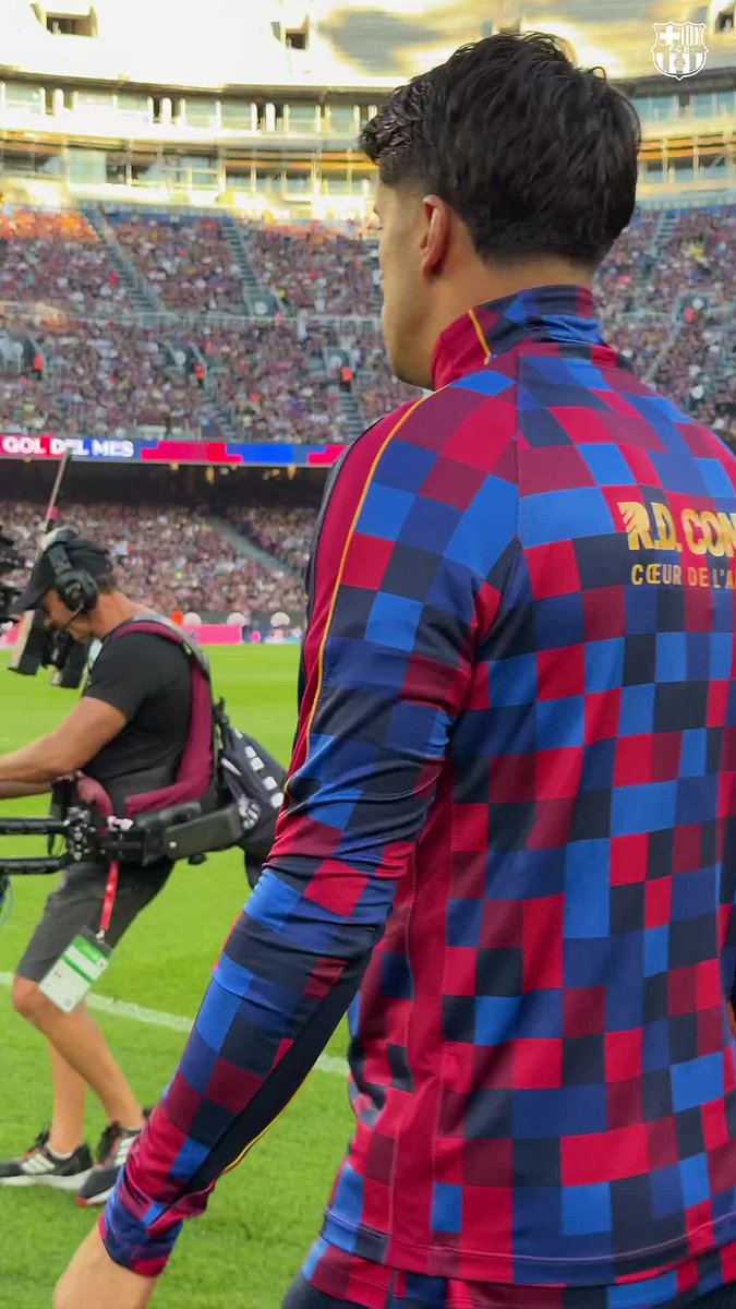 A footballer wearing a Barcelona pre-match top with a blue and red checkered pattern (text 'R.D. CON...', 'CŒUR DE L'...') walks on the pitch at Camp Nou while a cameraman films nearby.