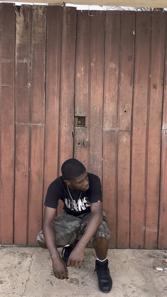 A young man wearing a black t-shirt, camouflage shorts, and black sneakers squats in front of a tall brown wooden door while looking to the side.
