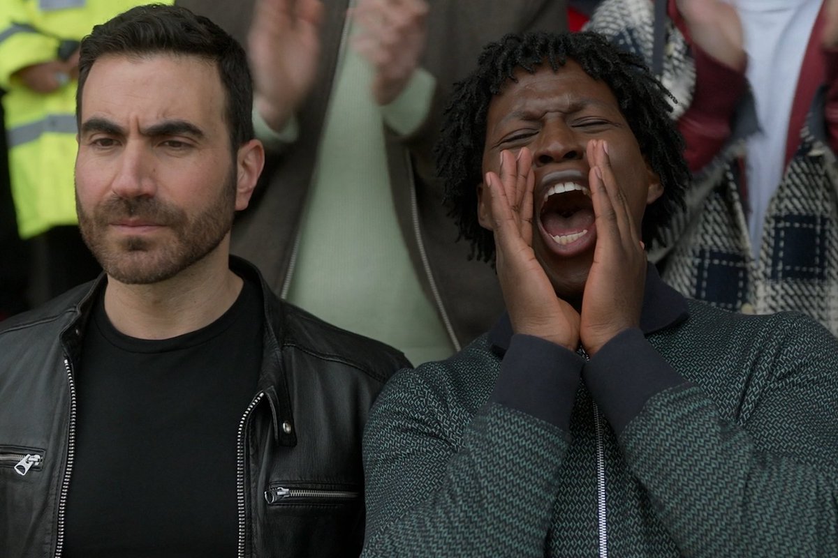 Two men sit in a stadium crowd, one stoic in a black leather jacket while the other enthusiastically shouts with hands cupped around his mouth, capturing the mixed emotions of devoted Richmond football fans in a scene from *TedLasso*.