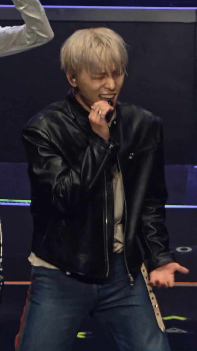 A performer with blond hair in a black leather jacket singing passionately into a microphone on a stage.