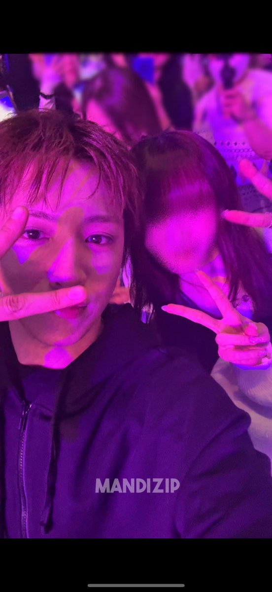 A selfie of a young man and woman at a concert bathed in pink and purple lighting, both flashing peace signs, with the watermark 'MANDIZIP' at the bottom.