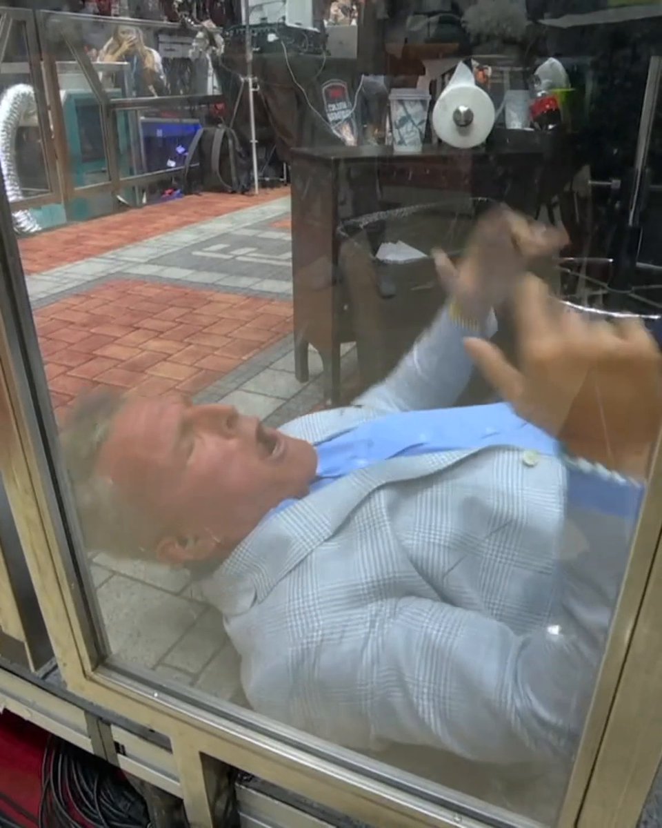 A humorous photo of a man in a light gray plaid blazer and blue shirt leaning back against a glass window with his mouth wide open in an exaggerated expression, while another person takes a picture from outside the glass.