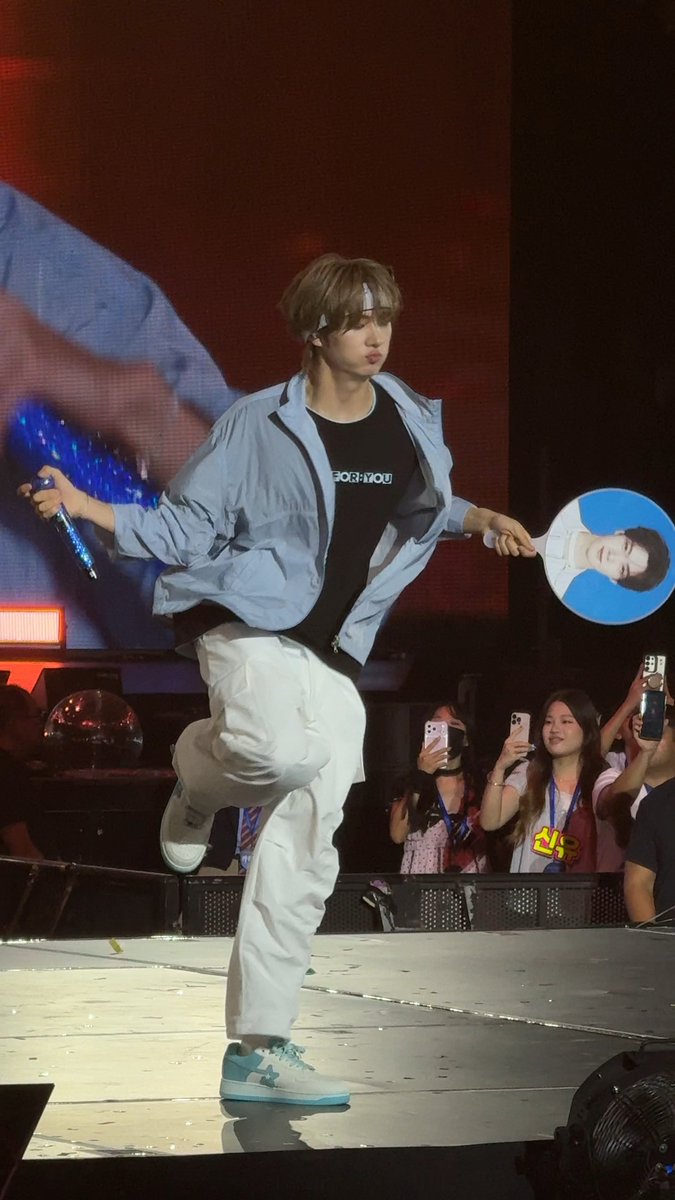 A performer on stage in a light blue jacket and white pants balances on one leg while holding a blue fan featuring a face, as fans in the background capture the moment on their phones.