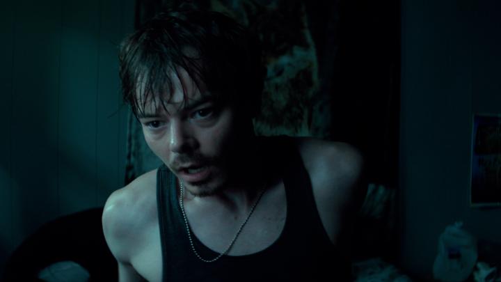 A young man with wet dark hair wearing a black tank top looks intensely off-camera in a dimly lit room with bluish lighting.