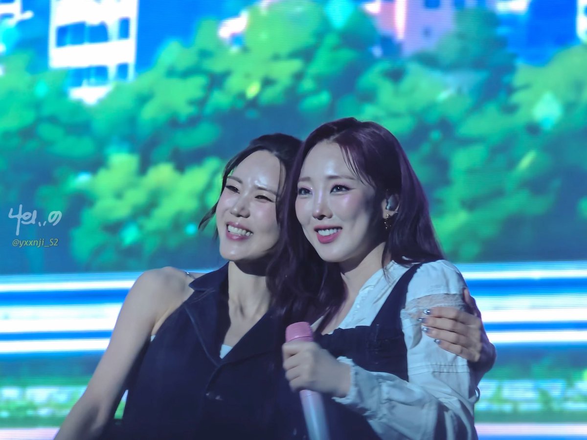 Two smiling women, identified by the watermark 'yxxnji_S2', embrace on stage in front of a large screen displaying a green forest scene; the text 'Yenny' is visible on the screen.