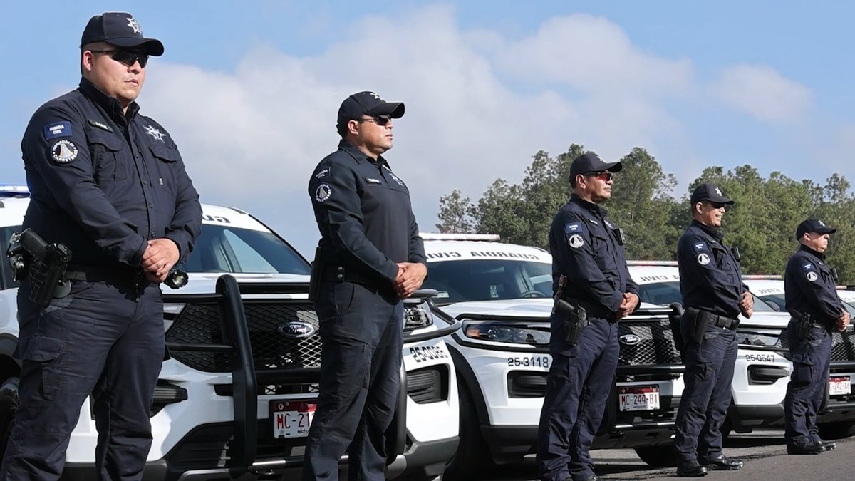 A line of uniformed police officers stands at attention in front of several white Ford patrol vehicles with license plates visible (MC-21, MC-244-B1). The vehicles have "SQUAD CANINO" written on them, indicating a canine unit.