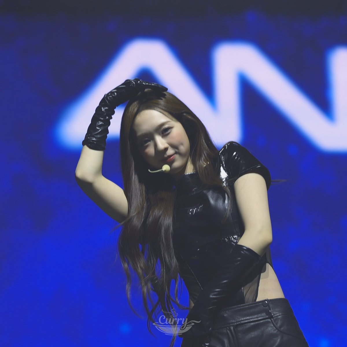 A young woman with long dark hair performing on stage in a black leather outfit and gloves, with blue lighting and a large 'M' logo in the background.