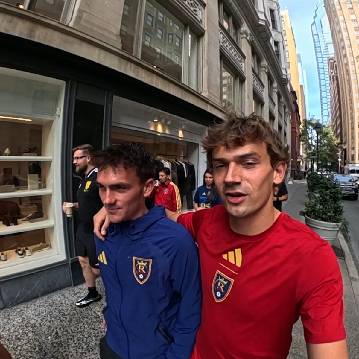 Two young men wearing Real Salt Lake soccer jerseys (one blue, one red) walk down a city sidewalk, with the man in red filming a selfie video.