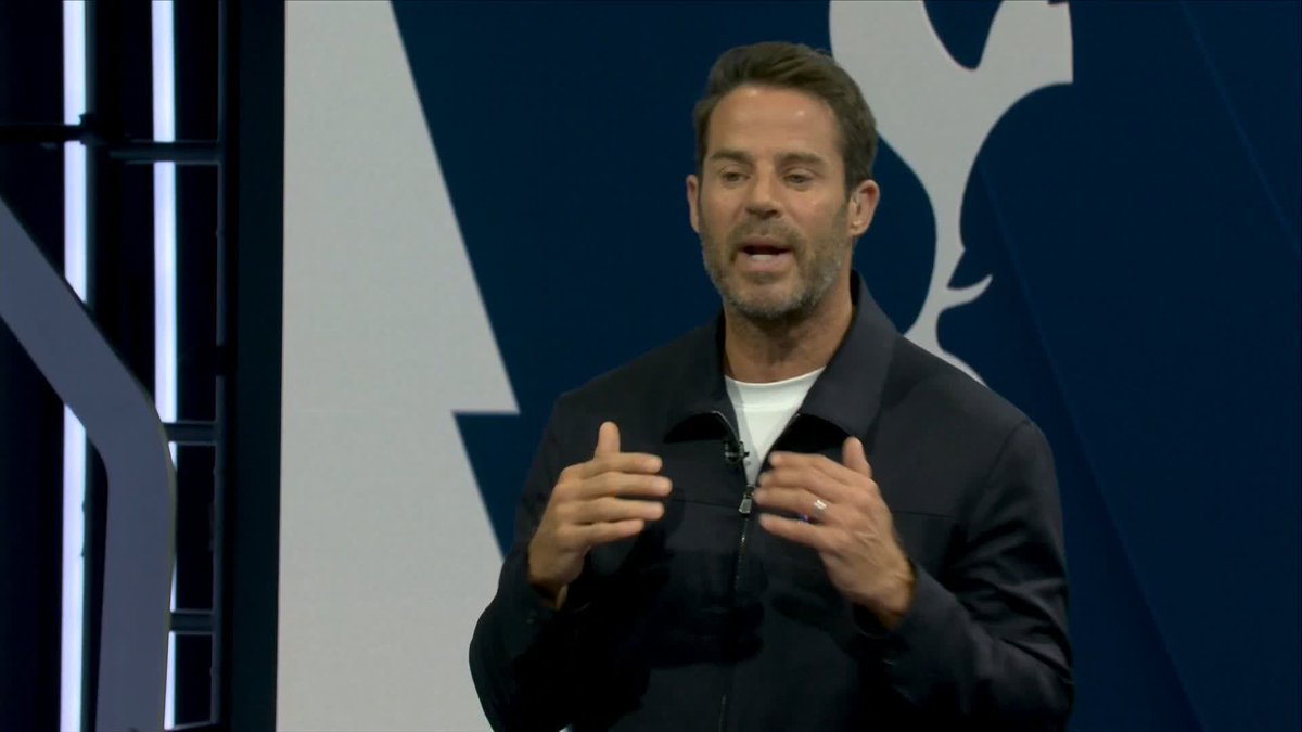 A man with short brown hair and a gray jacket over a white shirt is speaking and gesturing with his hands while standing in front of a blue and white patterned backdrop.