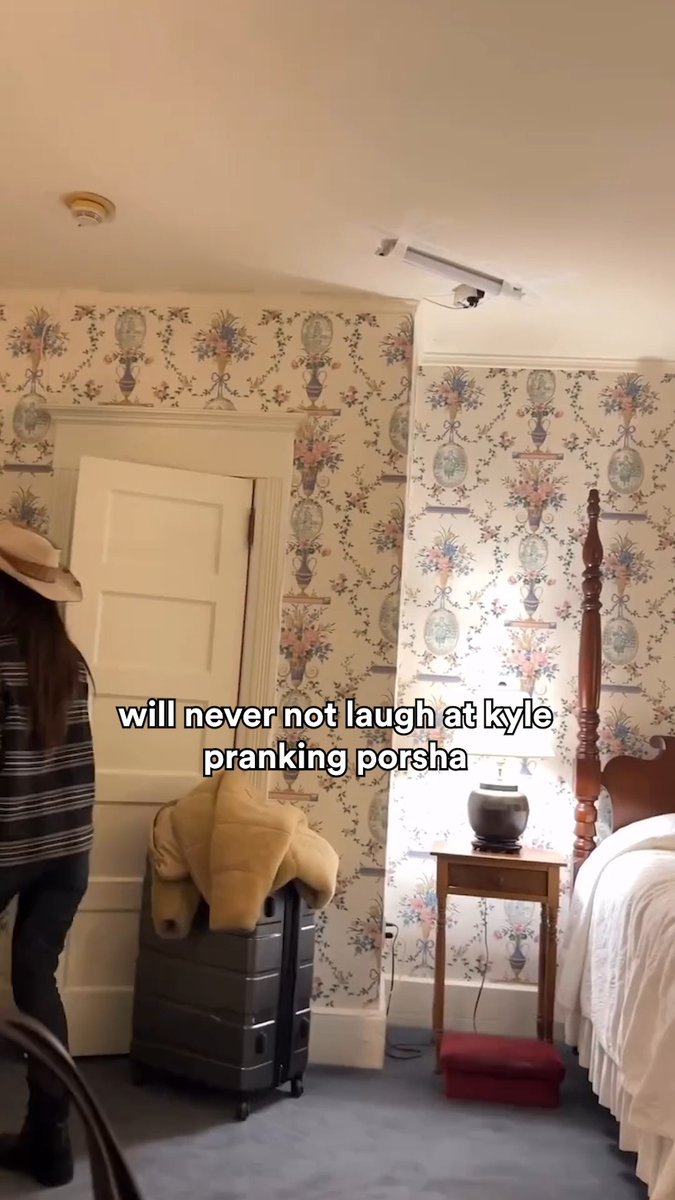 The image features a hotel room with floral wallpaper, where a person in a striped shirt and hat stands near a door, with a suitcase topped by what appears to be a horse-shaped pillow or costume on the floor. The on-screen text reads "will never not laugh at kyle pranking porsha," referencing a humorous moment from Real Housewives Ultimate Girls Trip that the post notes has been rewatched 500 times.