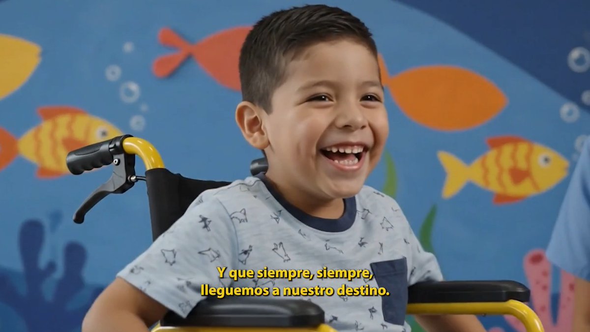 A young boy in a wheelchair smiles joyfully against a colorful fish-patterned wall, with Spanish subtitles reading 'Y que siempre, siempre, lleguemos a nuestro destino.'