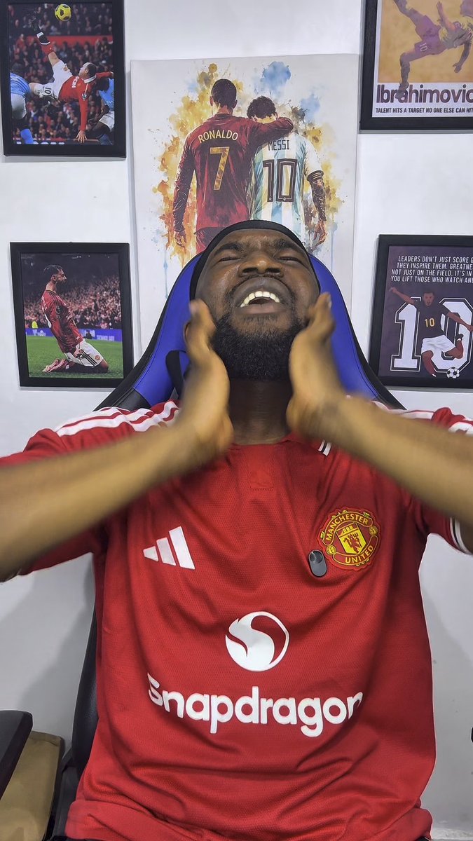 A man wearing a red Manchester United jersey with a Snapdragon sponsor logo sits in a blue and black gaming chair with his hands raised near his face and eyes closed, surrounded by framed soccer posters including Cristiano Ronaldo, Messi, and Ibrahimovic.