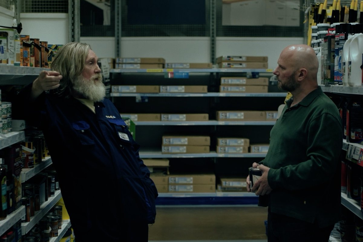 Two men converse in a dimly lit hardware or auto parts store aisle, surrounded by shelves stocked with boxed merchandise and bottles. One man, wearing a blue work jacket with a "TACKLE" name patch, gestures while speaking to a bald, bearded man in a green sweater holding a beverage can—evoking the modest, working-class setting fitting for the small-town preacher story of *Small Prophets*, now streaming on Apple TV.