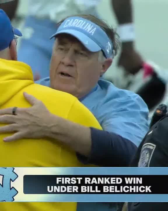 A man wearing a light blue "CAROLINA" visor and matching shirt embraces someone in a yellow hoodie, with on-screen text reading "FIRST RANKED WIN UNDER BILL BELICHICK." The image captures the moment celebrating Belichick's first victory over a ranked opponent as North Carolina's head coach, shown against the backdrop of a Pittsburgh crowd.
