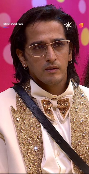A portrait of a man with shoulder-length dark hair wearing tinted glasses and an ornate white suit with gold sequined lapels and a bow tie, with 'BIGG BOSS S20' text visible in the corner.