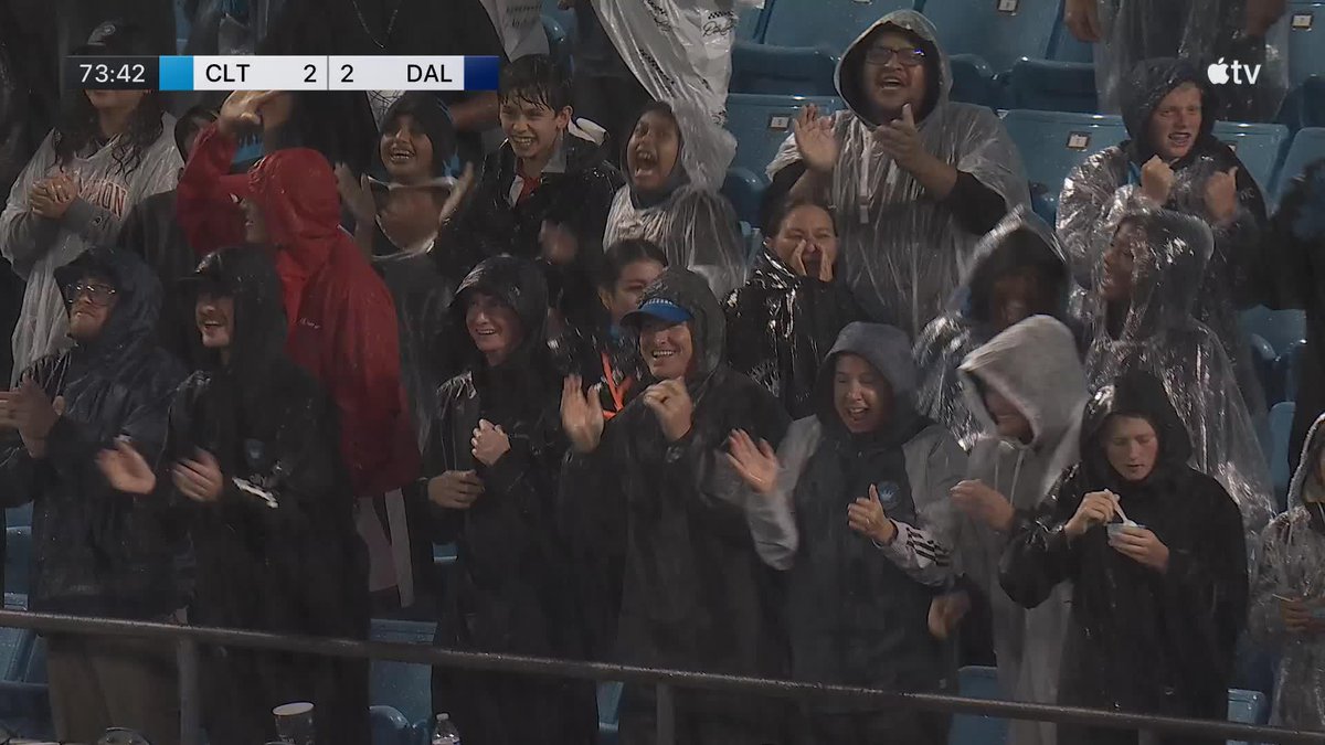 Fans in rain ponchos cheer enthusiastically at a Charlotte FC soccer match, where the scoreboard shows CLT 2, DAL 2 at the 73:42 mark, celebrating the equalizing goal mentioned in the post.
