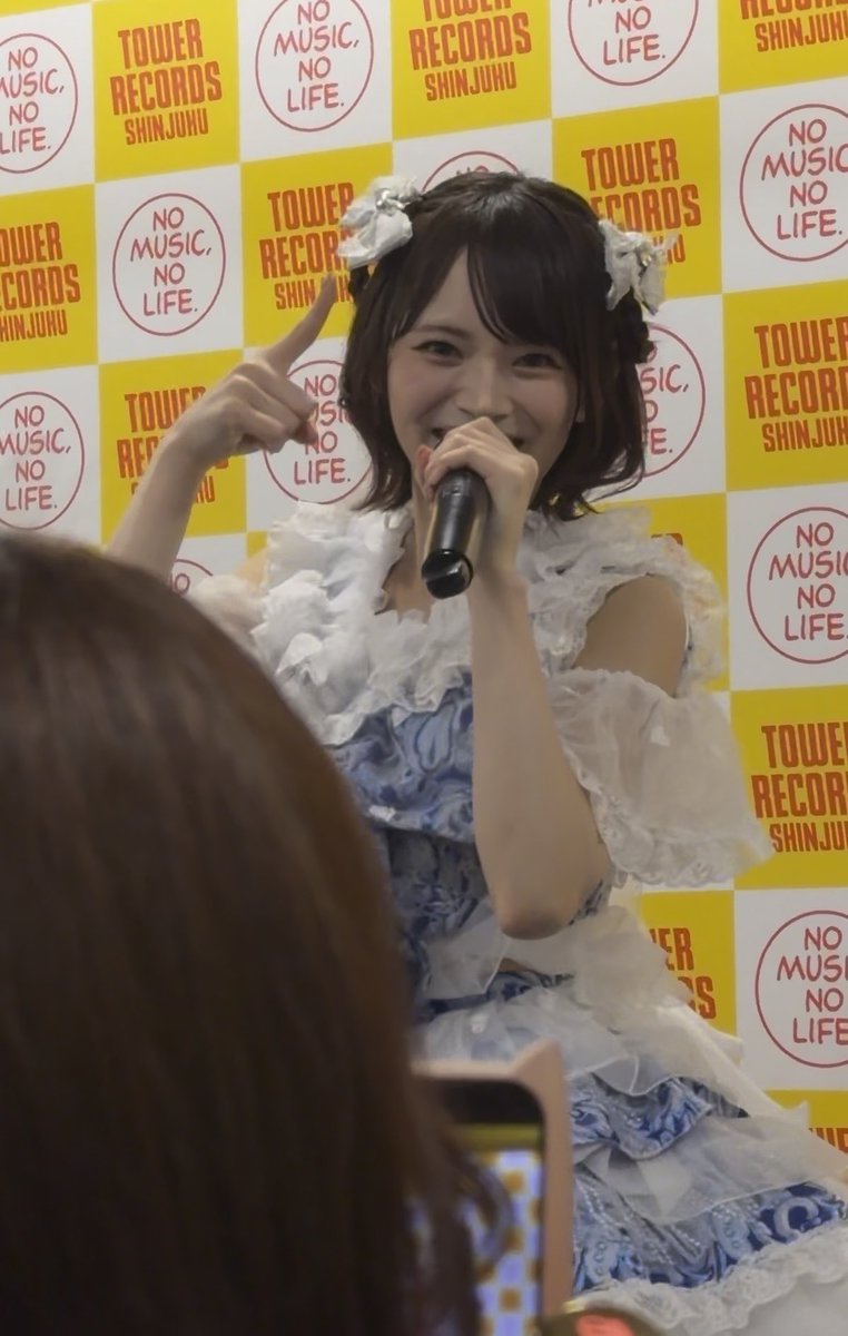A young woman with short brown hair and white flower hair clips, wearing a blue and white frilly dress, points upward while smiling into a microphone. The background features a yellow and white checkered pattern with 'NO MUSIC, NO LIFE' and 'TOWER RECORDS SHINJUKU' logos.