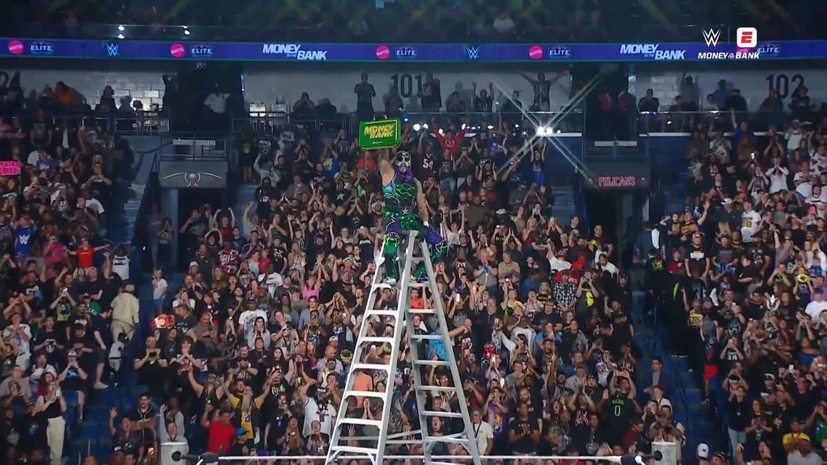 A WWE wrestling event where a masked performer in green and purple attire stands atop a ladder in the ring, holding a green 'Money in the Bank' briefcase above his head while the crowd cheers.