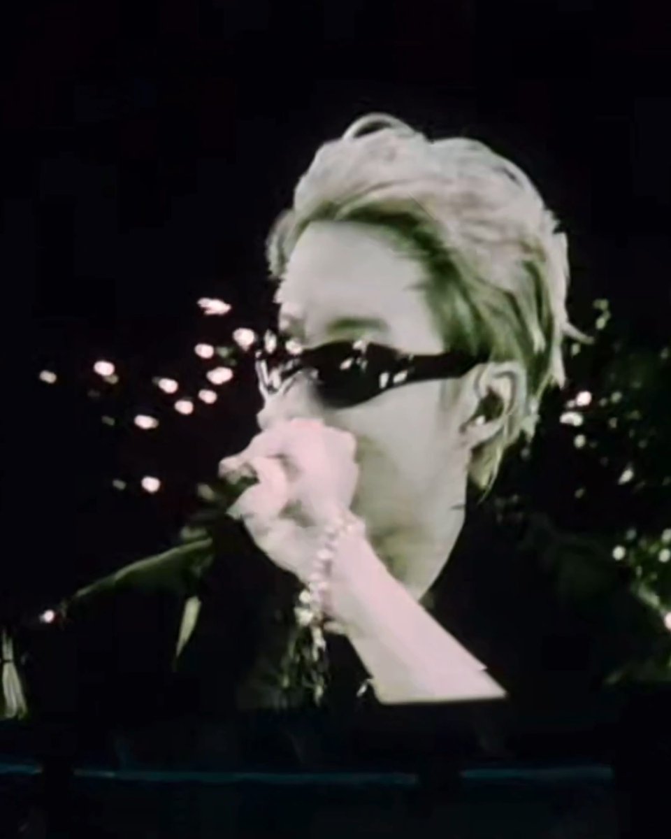 A person with light-colored hair wearing dark sunglasses speaks into a microphone at night, with bokeh lights in the background.