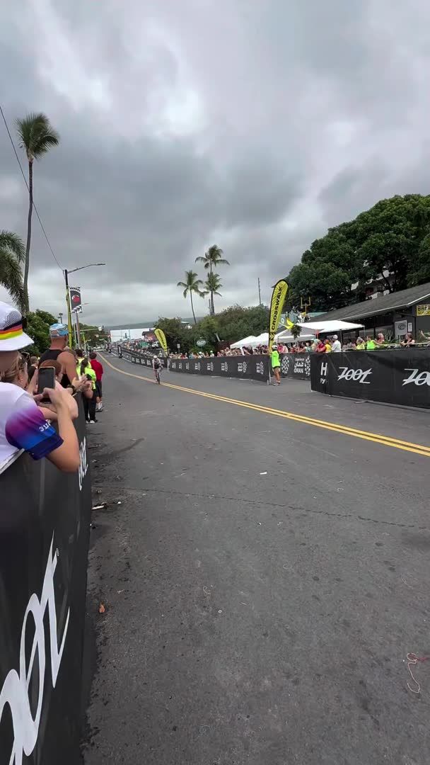 A street scene of a marathon race with runners on the course, spectators watching from behind barriers, palm trees, and a partly cloudy sky.