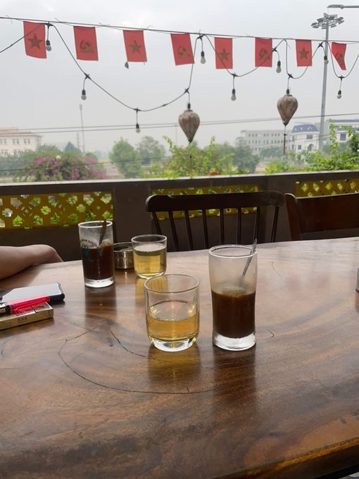 A wooden table on a balcony holds several glasses containing drinks, with strings lights and Vietnamese flags hanging above against a hazy outdoor backdrop.