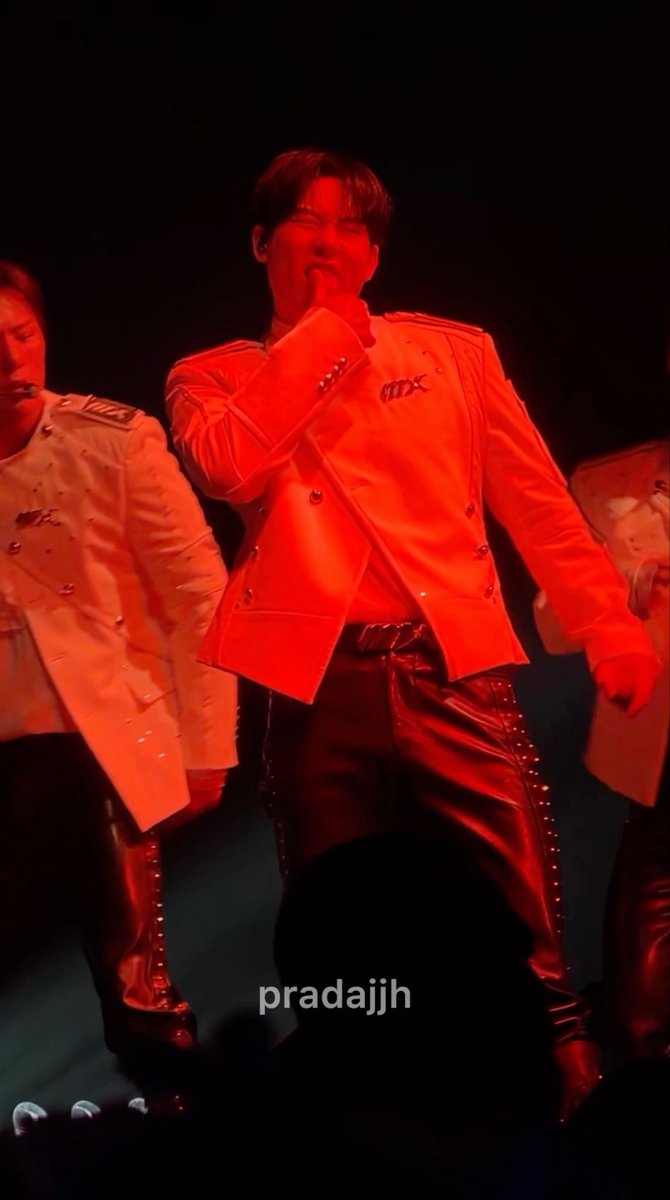 A performer in a white military-style jacket and black leather pants sings into a microphone under red stage lighting, with the watermark 'pradajjh' visible.