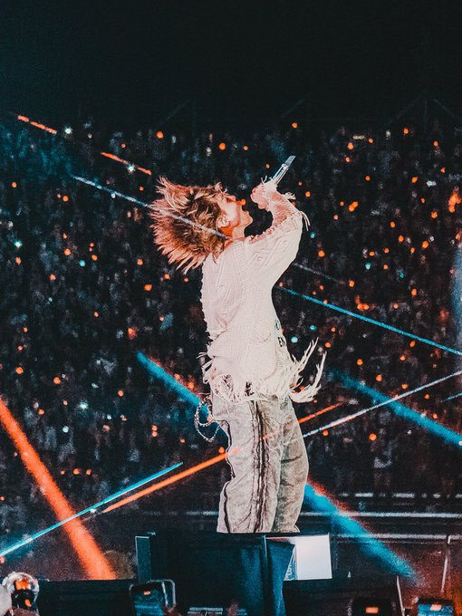 A performer with long reddish-brown hair stands on stage holding a microphone up to their mouth, wearing a white fringed shirt and patterned pants, with an orange light display and crowd lights visible behind them.