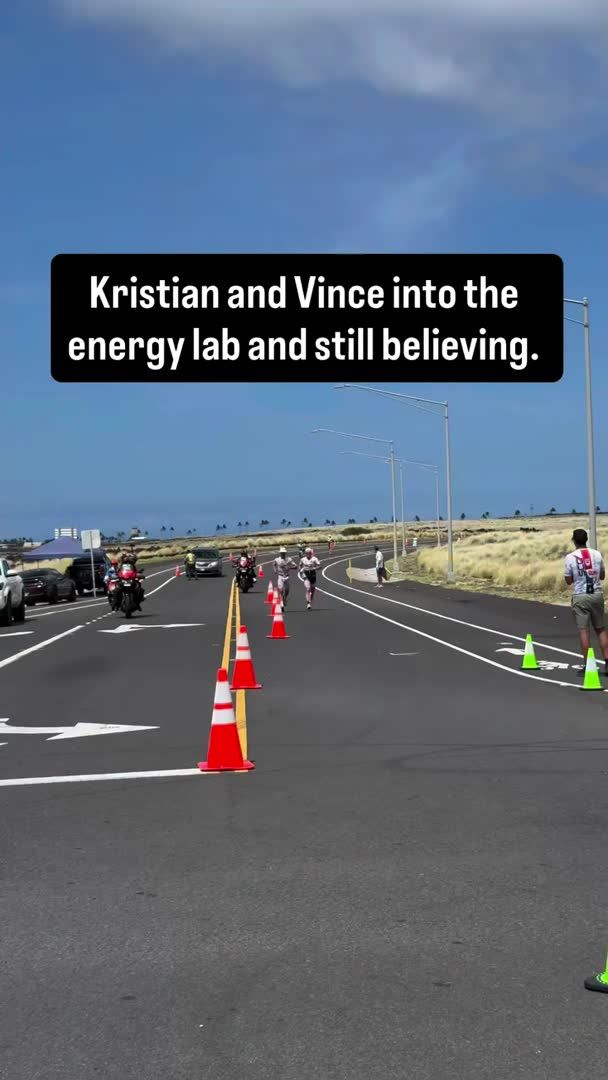 Outdoor shot of a road race with runners and motorcycles, with a black text overlay reading "Kristian and Vince into the energy lab and still believing."