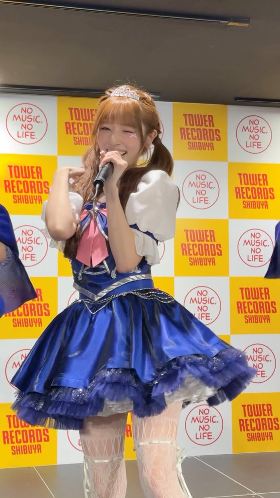A performer with a tiara and pigtails smiles while singing into a microphone at a Tower Records Shibuya event, wearing a blue and white dress with pink and silver accents. The background displays the iconic "Tower Records Shibuya" and "No Music, No Life." branding.