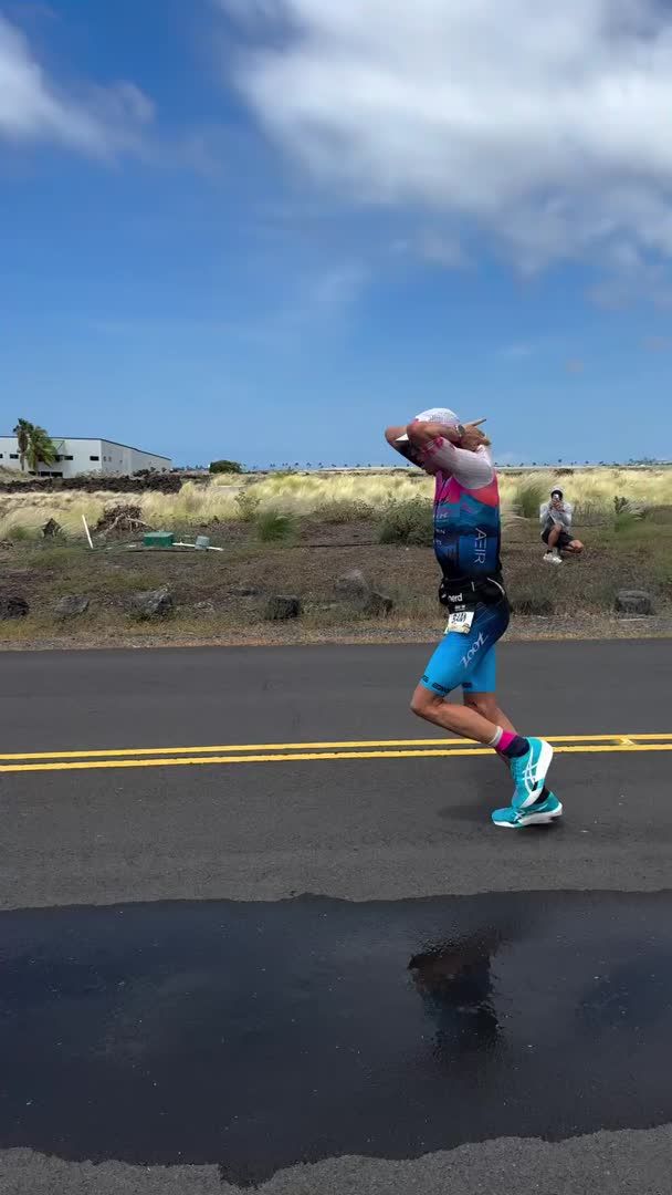 An athlete in a blue and pink triathlon suit runs on a wet road, appearing exhausted with hands raised near his head, while a person kneels in the background near grassy terrain.