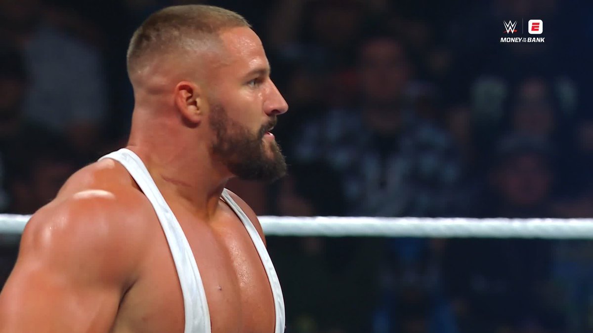 A muscular male wrestler with a short beard and buzz cut, wearing a white singlet, shown in profile at a WWE Money in the Bank event.