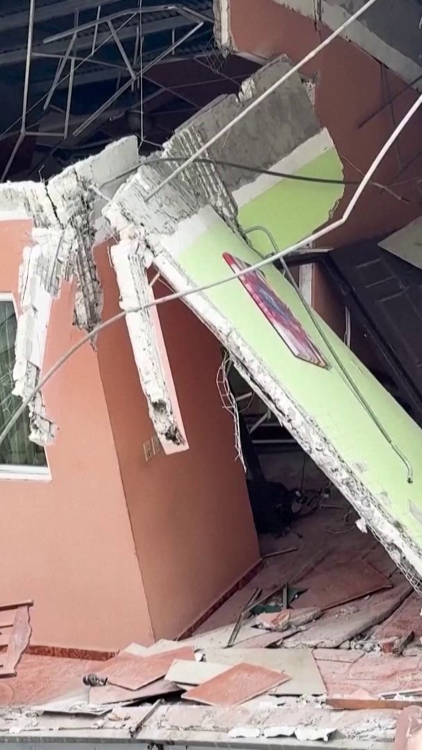 A photo of severe structural damage to a building, showing a collapsed concrete slab hanging down, exposed rebar, and debris scattered across a tiled floor.