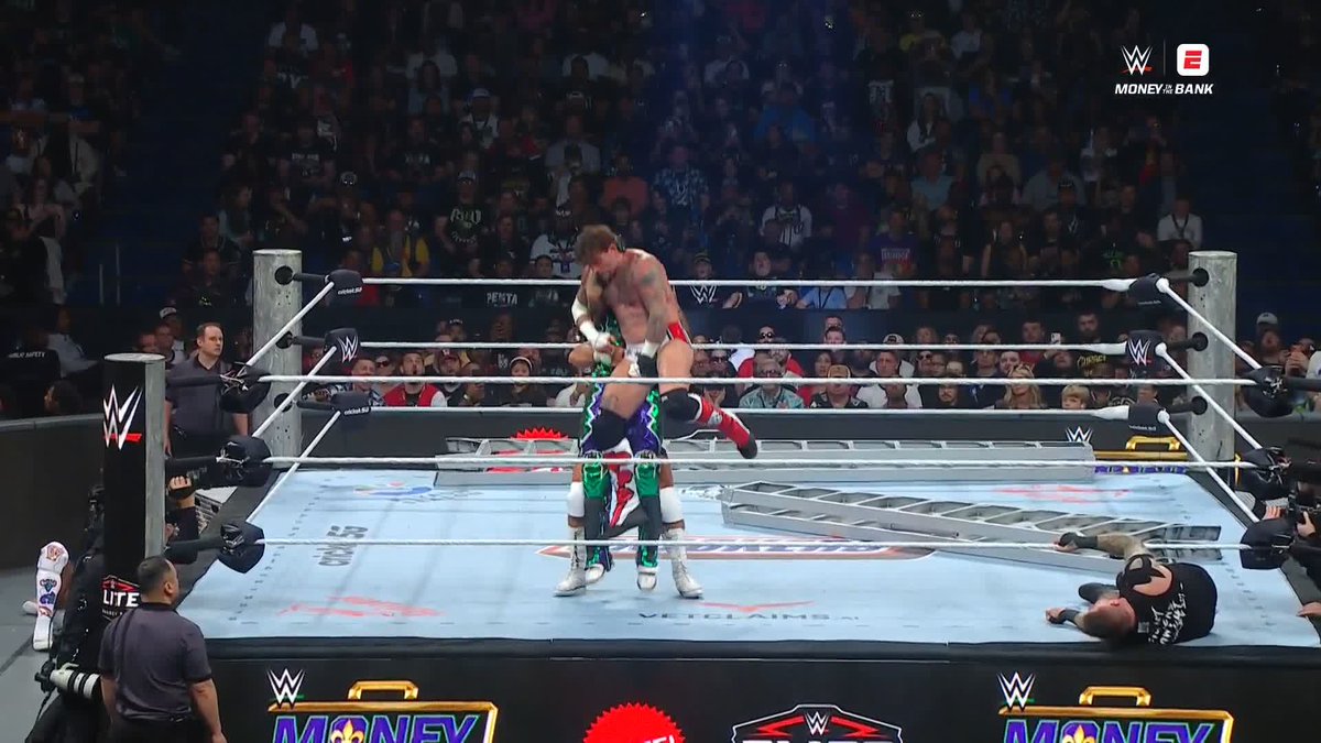 A WWE Money in the Bank wrestling match shows two wrestlers grappling in the ring while a referee lies on the mat and a ladder is set up nearby.