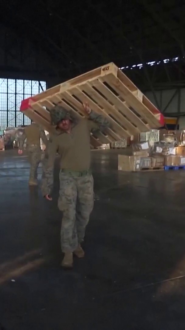A U.S. service member in camouflage uniform carries a large wooden pallet across their shoulders inside what appears to be a warehouse filled with supply crates, supporting U.S. Southern Command's humanitarian aid delivery efforts to Panama following the earthquakes. The pallet bears the marking "KY" in red lettering on its side.