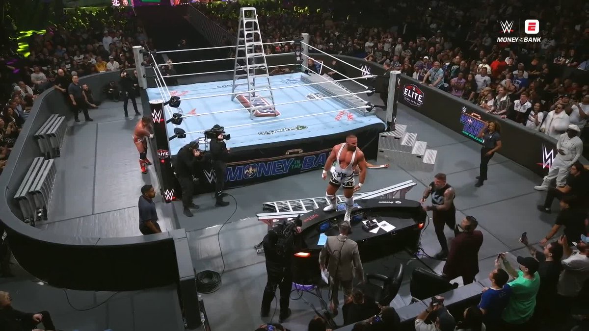 A WWE wrestling event scene showing wrestlers in and around a ring during a Money in the Bank match, with a ladder set up in the ring and the announce table destroyed in the foreground.