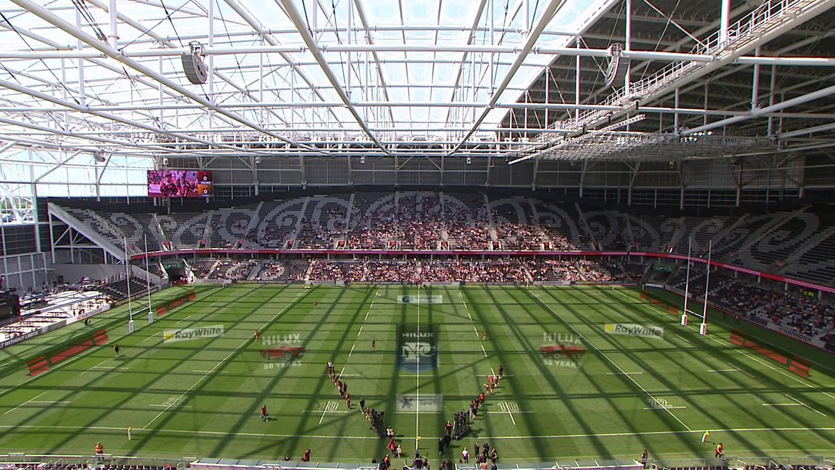 Wide interior view of a large stadium with a glass roof, showing a rugby field with players lined up, surrounded by advertising boards including 'RayWhite' and 'HILUX 50 YEARS'.