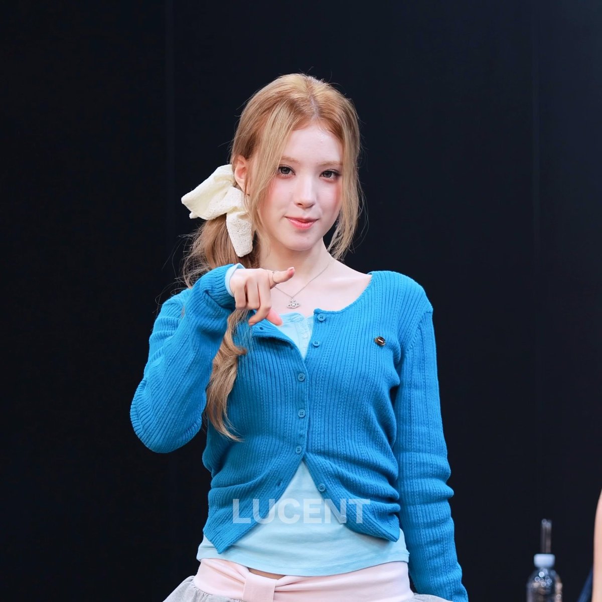 A young woman with blonde hair tied with a cream bow, wearing a blue cardigan, points at the camera in a fan event photo watermarked 'LUCENT'.