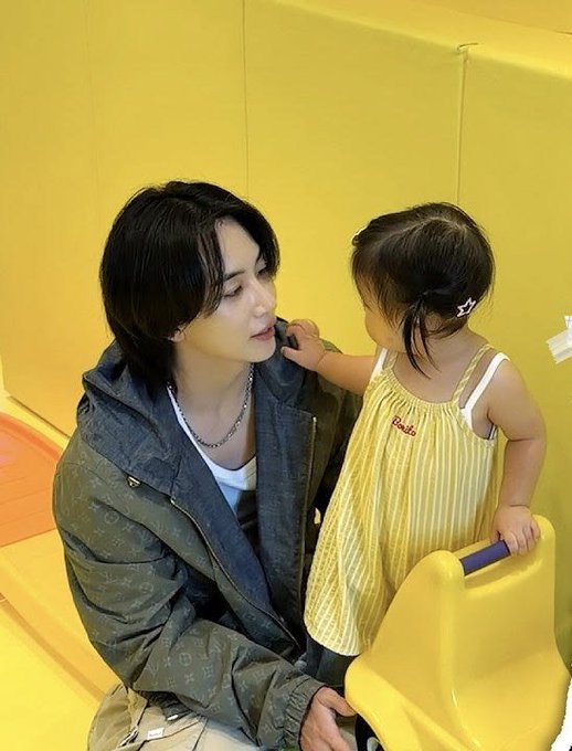 A man with dark hair wearing a monogrammed jacket sits next to a toddler in a yellow striped dress, interacting affectionately in what appears to be a yellow playroom.