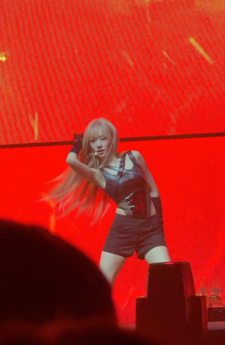 A performer with long blonde hair dances on stage in a black leather outfit against a bright red backdrop.