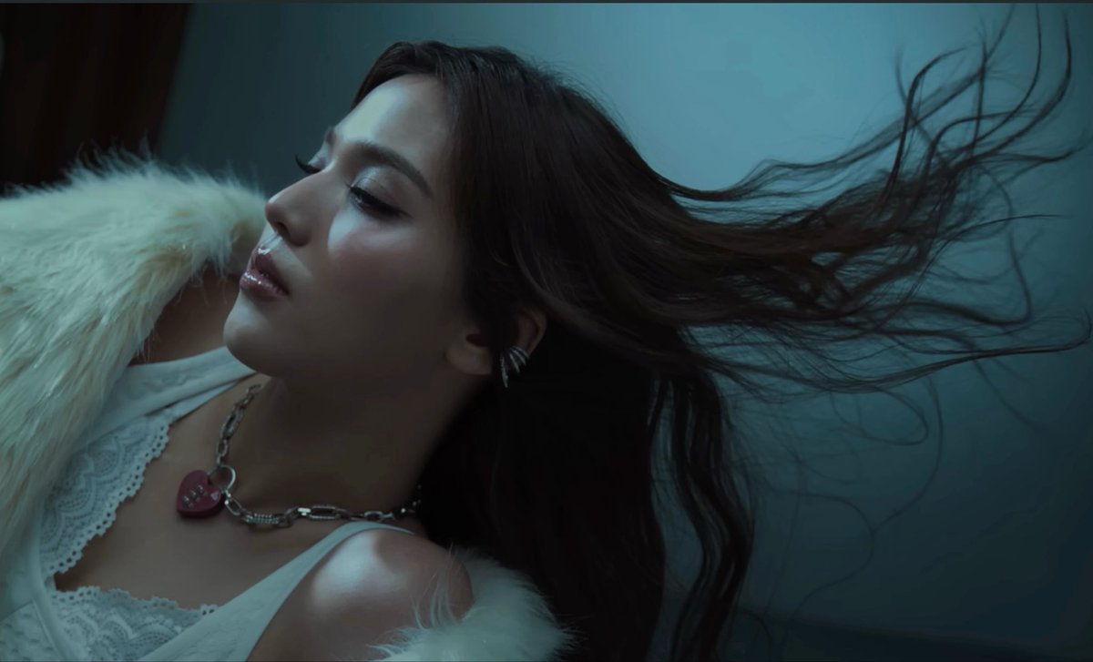 A woman lies with her eyes closed, her long dark hair dramatically swept upward against a blue-tinted background, wearing a white lace top, fluffy white garment, and a chain necklace with a red heart pendant.