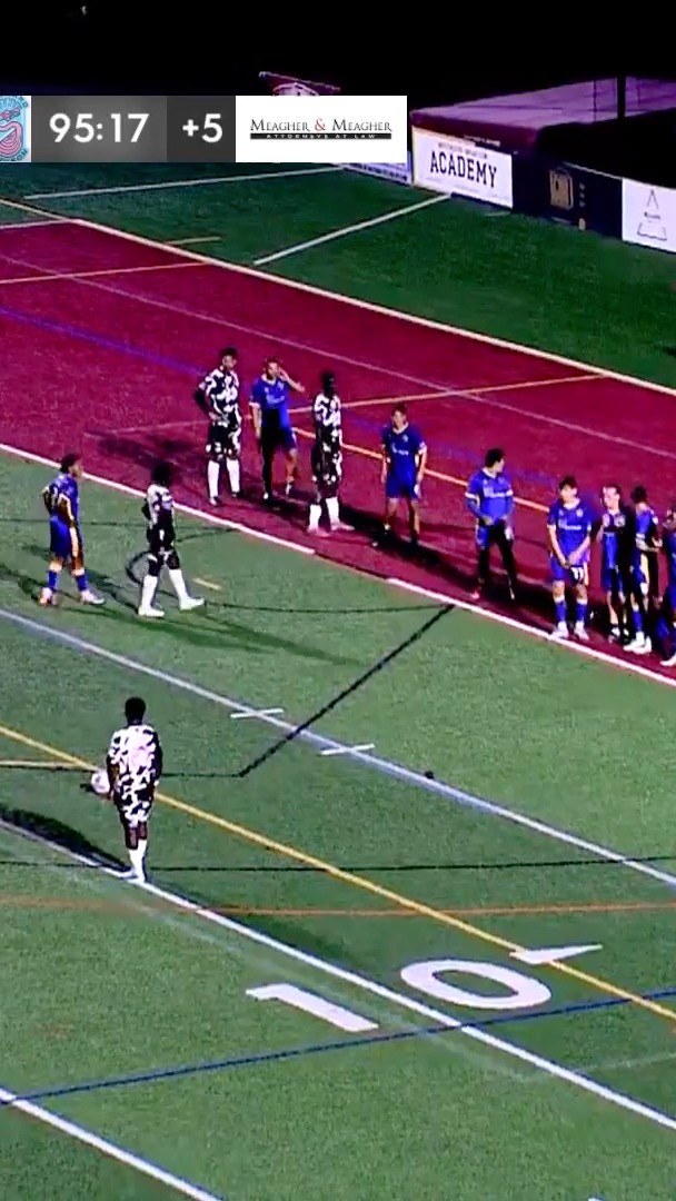 A nighttime soccer match shows players in blue and black-and-white cow-print uniforms lined up on the field, with a scoreboard displaying '95:17 +5' and banners for 'Meagher & Meagher' and 'ACADEMY'.