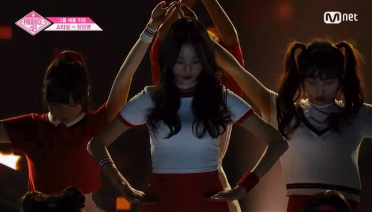 A screenshot from the Mnet show "Produce 48" showing three young female performers in cheerleader-style outfits (white tops with red accents) dancing on a dimly lit stage, with on-screen text reading "PRODUCE 48," "그룹 배틀 창단," "스타쉽 - 장원영," and the Mnet logo.