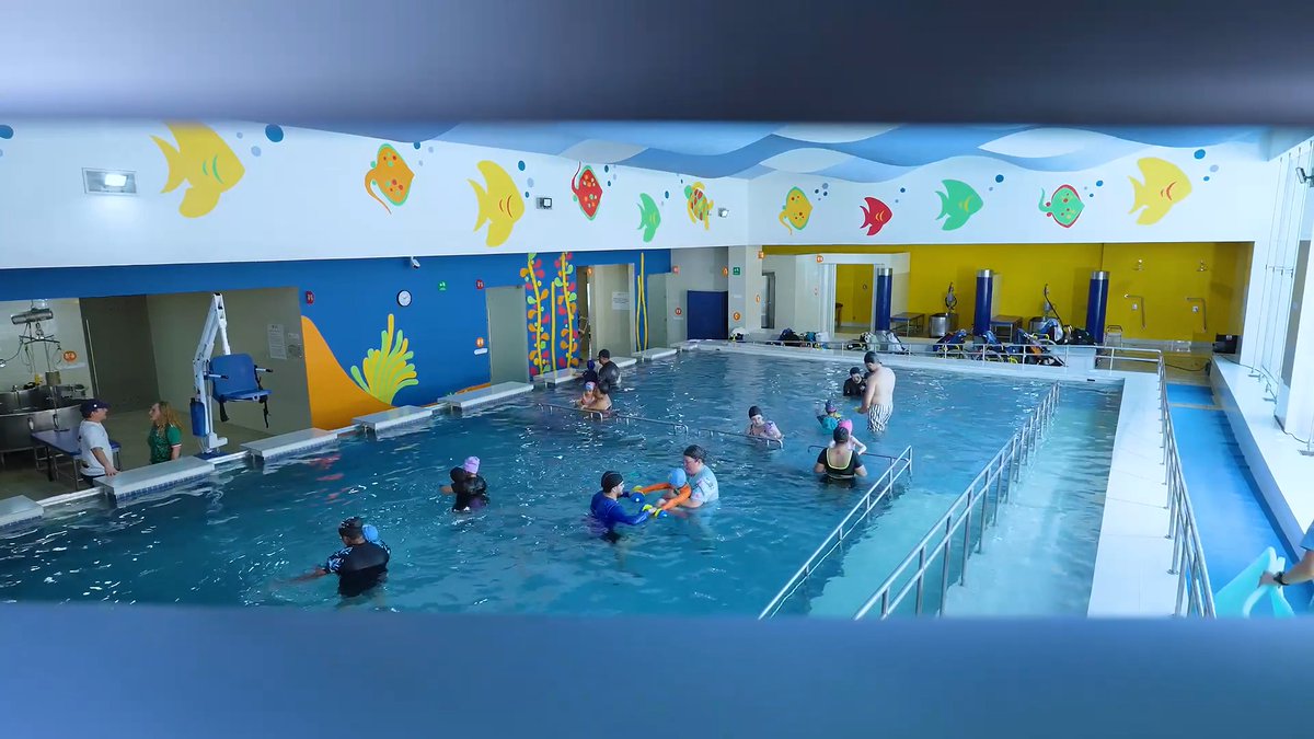An indoor swimming pool facility with colorful tropical fish and coral wall murals, where several children and adults are engaged in swimming lessons or aquatic activities, with a pool lift visible on the side.
